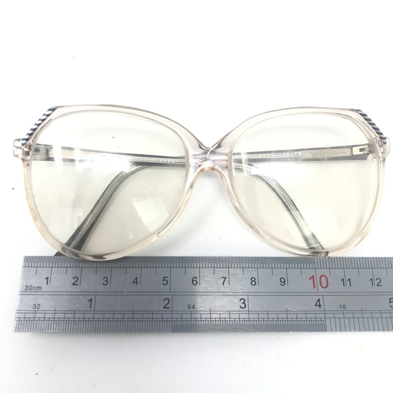 May include: Clear, oversized eyeglasses with a light-colored frame and dark temples. The glasses are placed on a white surface next to a ruler. The ruler shows measurements in inches and centimeters.