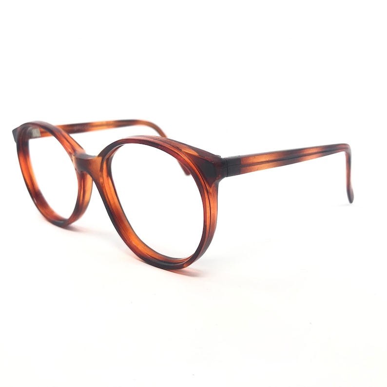May include: Round-framed eyeglasses in a tortoiseshell pattern. The glasses have a thick, dark brown frame with a lighter brown pattern. The lenses are clear, and the arms are thin and straight, with a slight curve at the end. The glasses are set against a white background.