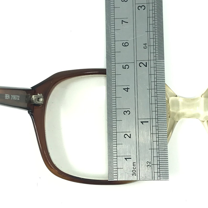 May include: Brown rectangular eyeglasses with clear lenses are measured with a ruler. The ruler shows a measurement of approximately 4 inches. The glasses have the text "BEN 21063732" on the arm.