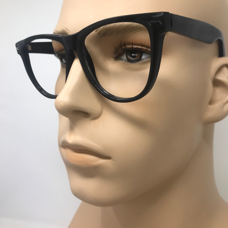 May include: Black-framed eyeglasses with a classic, slightly oversized design. The glasses have a thick, rectangular frame and clear lenses. The eyeglasses are displayed on a mannequin head against a white background.