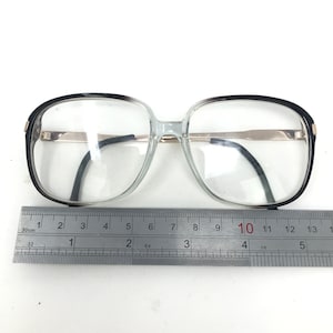 May include: Clear eyeglasses with a black and silver frame. The glasses have a rectangular shape and are measured with a ruler. The frame measures approximately 13 cm wide.