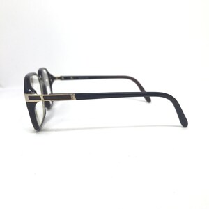 May include: Black-framed eyeglasses with a rectangular shape and gold detailing. The lenses are clear, and the arms are dark brown. The glasses are shown against a white backdrop.