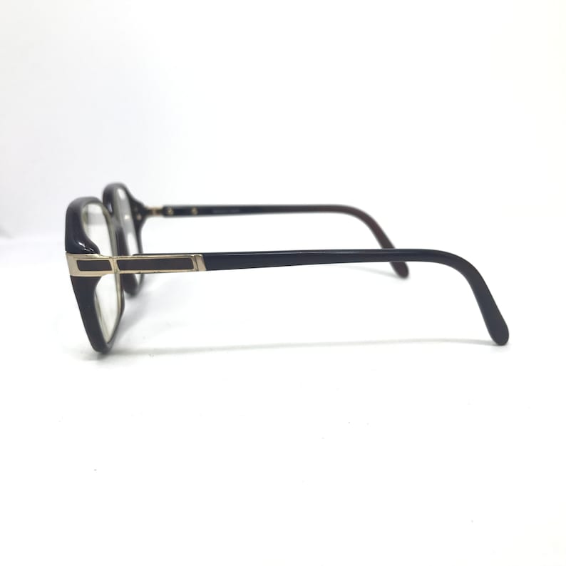 May include: Black-framed eyeglasses with a rectangular shape and gold detailing. The lenses are clear, and the arms are dark brown. The glasses are shown against a white backdrop.