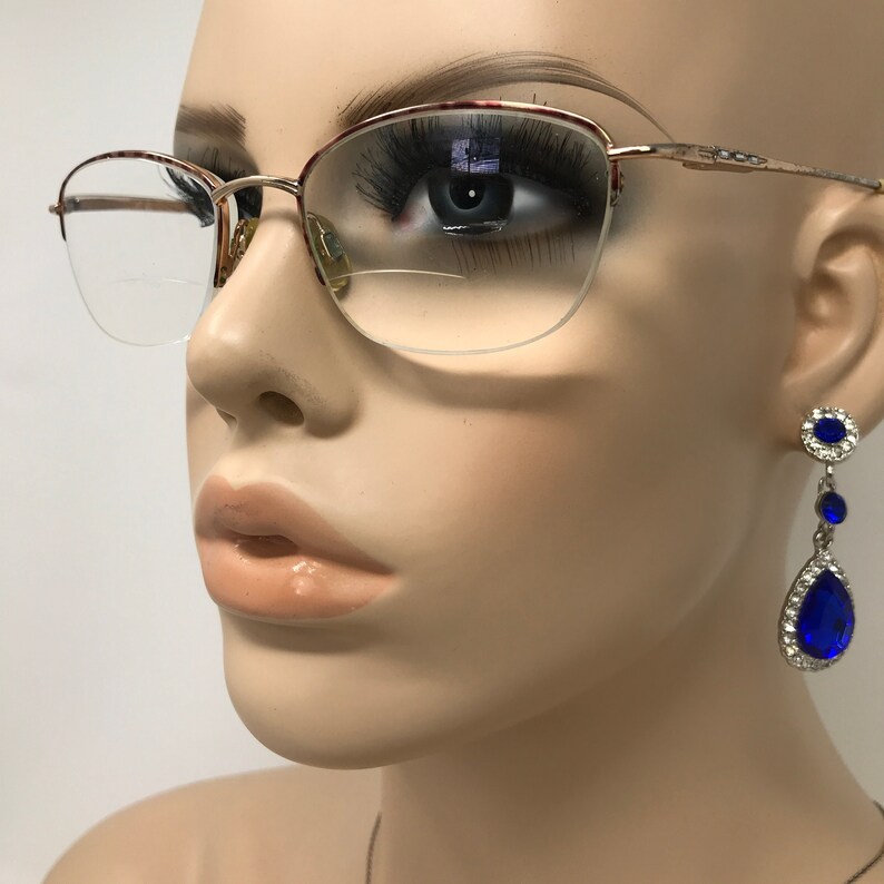 May include: Gold-rimmed eyeglasses with a half-frame design and a tortoiseshell pattern on the upper rims. The image also features a pair of dangling blue teardrop earrings with a silver setting, showcasing a glamorous aesthetic.