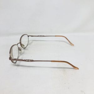 Op de afbeelding: Een bril van bruin metaal met ovale glazen. De pootjes van de bril zijn in een lichtere bruine tint.