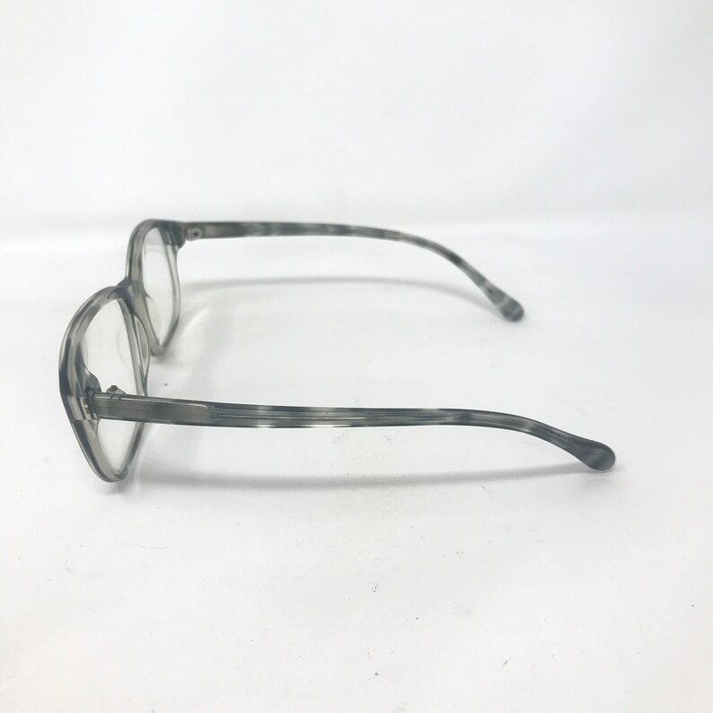 Puede incluir: Gafas grises con un patr&oacute;n veteado. Las monturas son de color gris claro y las lentes son transparentes. Las patillas de las gafas se curvan suavemente para adaptarse a las orejas. Las gafas est&aacute;n sobre un fondo blanco.