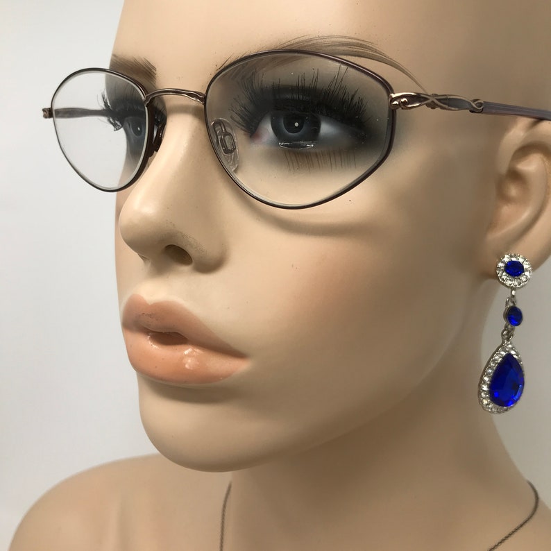 Puede incluir: Maniqu&iacute; con gafas ovaladas con montura fina y oscura. El maniqu&iacute; tambi&eacute;n lleva un par de pendientes colgantes con gemas azules en forma de l&aacute;grima y gemas azules m&aacute;s peque&ntilde;as rodeadas de piedras transparentes.