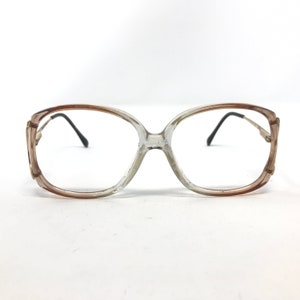 May include: Vintage eyeglasses with a clear and brown gradient frame. The glasses have a large, rounded shape with black temples and small gold accents on the corners. The lenses are clear.