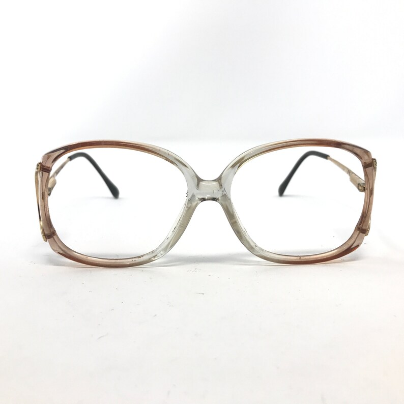 May include: Vintage eyeglasses with a clear and brown gradient frame. The glasses have a large, rounded shape with black temples and small gold accents on the corners. The lenses are clear.