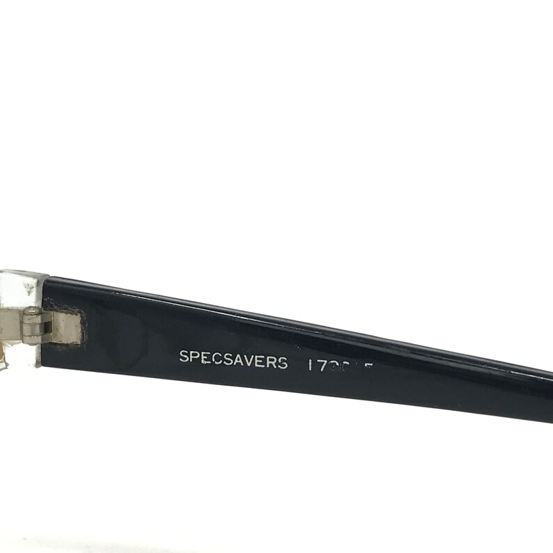 May include: Close-up of a black eyeglass frame arm with the text "SPECSAVERS" in white. The frame is black with a silver hinge and a clear plastic section near the lens.