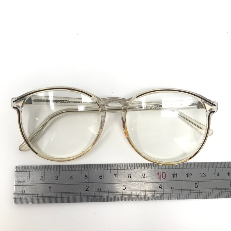 Puede incluir: Gafas transparentes con montura redonda y una regla para medir. Las gafas tienen una montura transparente con un ligero tinte dorado alrededor de las lentes. El texto "MC-9205" es visible en la montura. La regla muestra medidas en cent&iacute;metros.
