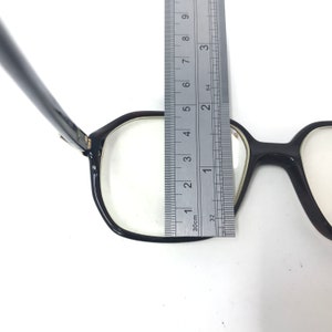 May include: Black-framed eyeglasses with clear lenses. A ruler measures the frame, showing a width of approximately 5 cm. The glasses have a classic, rectangular shape and are set against a white background.