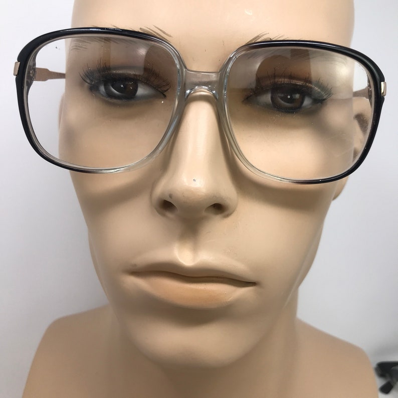 May include: Eyeglasses with clear frames, black upper rims, and gold-coloured accents. The square lenses are set in a clear plastic frame, with a black border on the top and sides. The temples are gold-coloured.