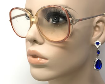 May include: A pair of large, round, brown-rimmed spectacles with clear lenses. The glasses are worn by a mannequin with blue teardrop earrings.
