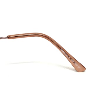 May include: Close-up of the temple arm of eyeglasses. The arm is a warm rose gold color and features the text "Specsavers 1001069 ce" printed in white. The arm curves gently and tapers towards the end.