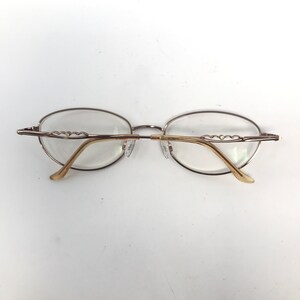 May include: Oval-shaped eyeglasses with a gold-colored metal frame. The temples have a decorative wave design. The lenses are clear. The glasses are set against a white background.
