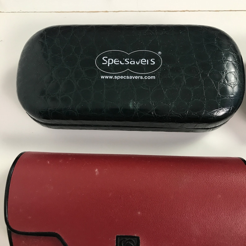 May include: Two eyeglass cases. The top case is black with a textured, crocodile-like pattern and the Specsavers logo. The bottom case is burgundy with a smooth finish and a black clasp.