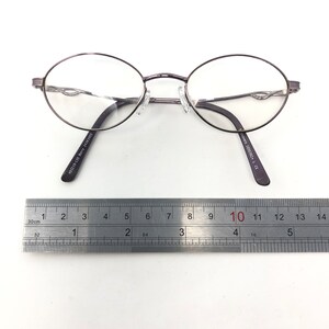 Peut inclure: Lunettes de vue ovales avec une monture en m&eacute;tal gris fonc&eacute;. Les lunettes ont des verres transparents et un pont de nez avec des plaquettes r&eacute;glables. Les branches sont gris fonc&eacute; avec le texte "49015-135 Berry 24383905" et "csavers 1000382+ L C". Une r&egrave;gle est plac&eacute;e en dessous.