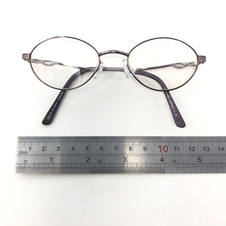 Peut inclure: Lunettes de vue ovales avec une monture en m&eacute;tal gris fonc&eacute;. Les lunettes ont des verres transparents et un pont de nez avec des plaquettes r&eacute;glables. Les branches sont gris fonc&eacute; avec le texte "49015-135 Berry 24383905" et "csavers 1000382+ L C". Une r&egrave;gle est plac&eacute;e en dessous.