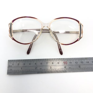 K&ouml;nnte beinhalten: Vintage Brille mit klaren Gl&auml;sern und einem silbernen Rahmen. Die Fassungen haben einen transparenten Kunststoffsteg und B&uuml;gel mit einem dunkelburgunderfarbenen und goldenen Akzent an den oberen R&auml;ndern. Ein Lineal ist darunter platziert.
