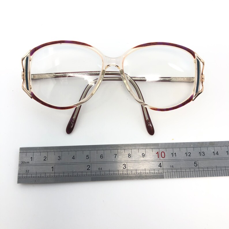 K&ouml;nnte beinhalten: Vintage Brille mit klaren Gl&auml;sern und einem silbernen Rahmen. Die Fassungen haben einen transparenten Kunststoffsteg und B&uuml;gel mit einem dunkelburgunderfarbenen und goldenen Akzent an den oberen R&auml;ndern. Ein Lineal ist darunter platziert.