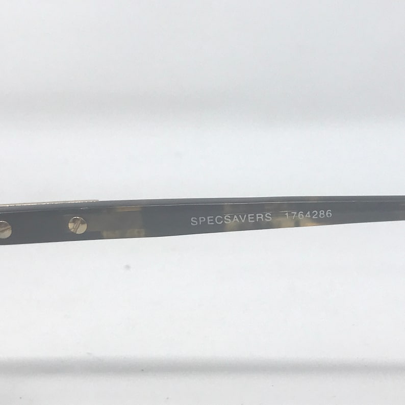 Puede incluir: Primer plano de gafas con montura oscura estampada. El texto "SPECSAVERS 1764286" est&aacute; impreso en la varilla. Las gafas est&aacute;n sobre un fondo blanco.
