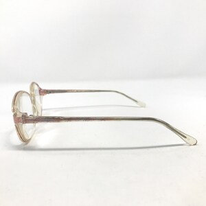 K&ouml;nnte beinhalten: Eine Brille mit runden, transparenten Gl&auml;sern und einem rosafarbenen und silbernen Design. Die Brille ist von der Seite zu sehen, wobei der rechte B&uuml;gel sichtbar ist.