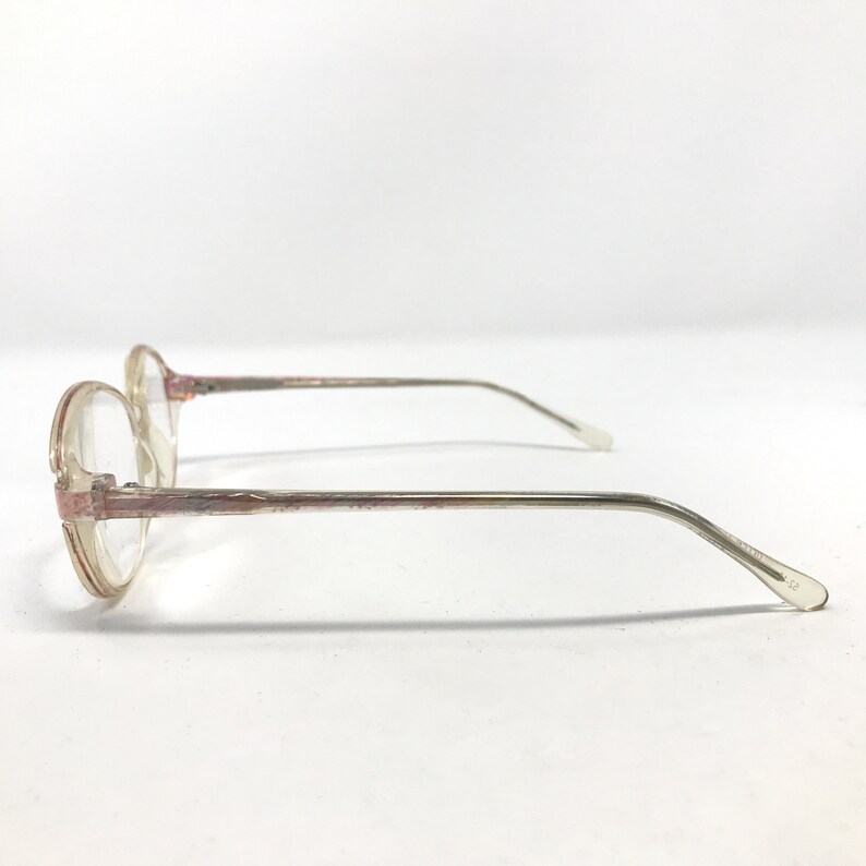 K&ouml;nnte beinhalten: Eine Brille mit runden, transparenten Gl&auml;sern und einem rosafarbenen und silbernen Design. Die Brille ist von der Seite zu sehen, wobei der rechte B&uuml;gel sichtbar ist.