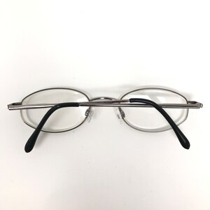 Op de afbeelding: Bril met zilveren montuur, ovale glazen en zwarte pootjes. De bril heeft een minimalistisch design, met een dun metalen montuur en heldere glazen. De neuspads zijn transparant. Geschikt voor gezichtscorrectie.