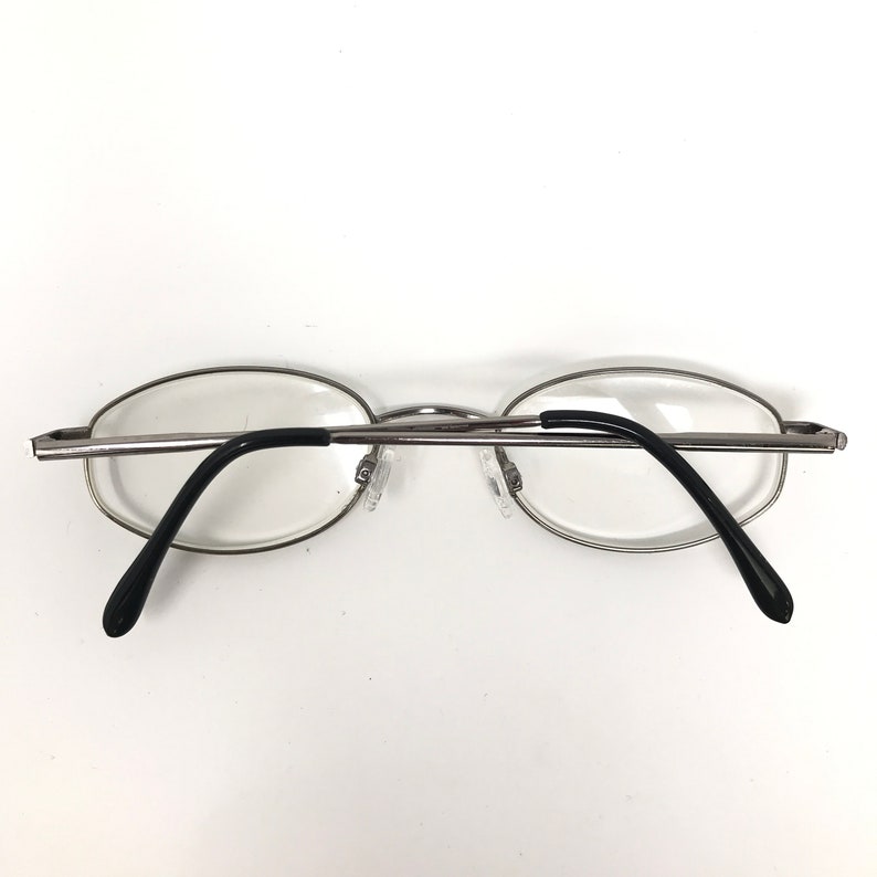 Op de afbeelding: Bril met zilveren montuur, ovale glazen en zwarte pootjes. De bril heeft een minimalistisch design, met een dun metalen montuur en heldere glazen. De neuspads zijn transparant. Geschikt voor gezichtscorrectie.