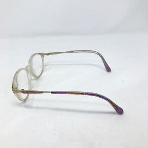 May include: Clear-framed eyeglasses with thin, silver-toned metal arms. The earpieces are accented with purple and gold detailing. The lenses are round, and the glasses are set against a white background.
