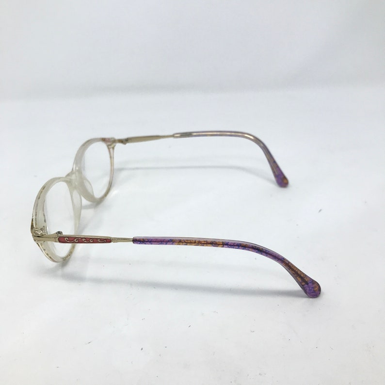May include: Clear-framed eyeglasses with thin, silver-toned metal arms. The earpieces are accented with purple and gold detailing. The lenses are round, and the glasses are set against a white background.