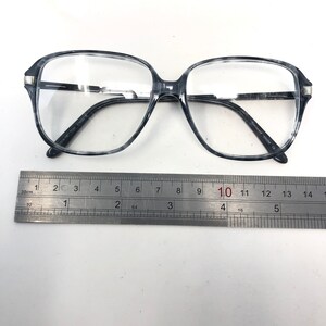 May include: Gray and white marbled eyeglasses with a rectangular shape. The glasses have clear lenses and a silver accent on the temples. A ruler is placed below the glasses, showing measurements in centimetres.