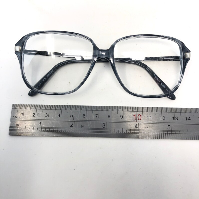May include: Gray and white marbled eyeglasses with a rectangular shape. The glasses have clear lenses and a silver accent on the temples. A ruler is placed below the glasses, showing measurements in centimetres.