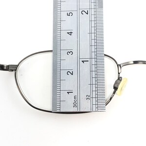 Puede incluir: Primer plano de gafas con montura de metal plateado. Una regla se coloca sobre la lente, midiendo aproximadamente 7,6 cm. Las gafas tienen una lente transparente y una almohadilla nasal.