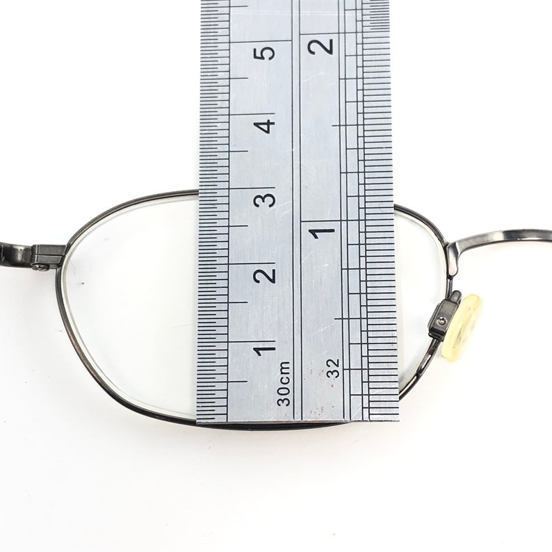 Puede incluir: Primer plano de gafas con montura de metal plateado. Una regla se coloca sobre la lente, midiendo aproximadamente 7,6 cm. Las gafas tienen una lente transparente y una almohadilla nasal.