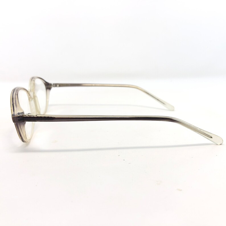 May include: A pair of eyeglasses with a clear, oval-shaped frame and thin, transparent arms. The lenses are clear, and the frame has a subtle, translucent gray hue. The glasses are designed for vision correction.