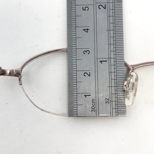May include: Close-up of eyeglasses with a silver frame. A ruler is placed next to the glasses, showing measurements in inches and centimeters. The glasses have clear lenses and a delicate design.