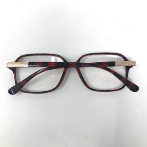 May include: Eyeglasses with a tortoiseshell pattern frame and clear lenses. The frame has a rectangular shape with a black and red pattern. The temples are black with a gold-colored accent at the hinge.