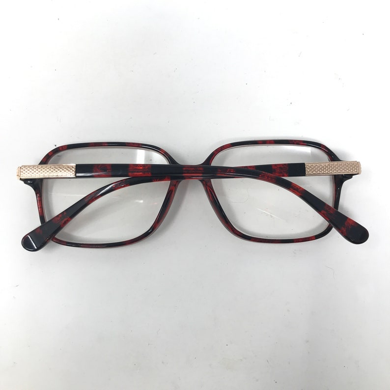 May include: Eyeglasses with a tortoiseshell pattern frame and clear lenses. The frame has a rectangular shape with a black and red pattern. The temples are black with a gold-colored accent at the hinge.