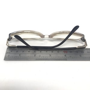 May include: Clear-framed eyeglasses with black temples, resting on a white surface. The glasses are positioned next to a ruler, with measurements in centimeters. The frame is a light, translucent color.