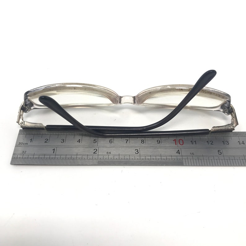 May include: Clear-framed eyeglasses with black temples, resting on a white surface. The glasses are positioned next to a ruler, with measurements in centimeters. The frame is a light, translucent color.