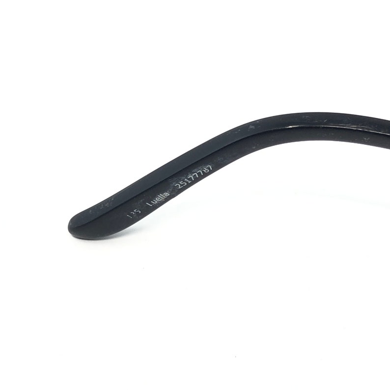 May include: Close-up of a black eyeglass arm. The text "135 Luella 25177787" is printed on the arm. The arm is curved and has a smooth finish. The background is white.