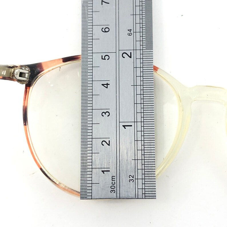 K&ouml;nnte beinhalten: Nahaufnahme einer Brille mit einem Lineal, das den Rahmen misst. Die Brille hat auf einer Seite ein Schildpattmuster und auf der anderen Seite einen klaren, hellfarbigen Rahmen. Das Lineal zeigt ein Ma&szlig; von ca. 3,8 cm.
