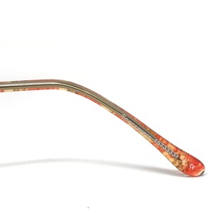 May include: Close-up of the arm of eyeglasses. The arm is gold-colored with a pattern of red, orange, and beige. The text "Specsavers 1573333+" is printed on the arm.