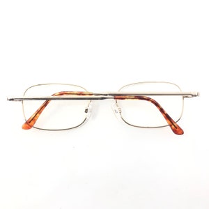 May include: Rimless spectacles with a gold-coloured metal frame and tortoiseshell patterned arms. The spectacles have a rectangular shape and clear lenses. The nose pads are clear. Designed for vision correction.