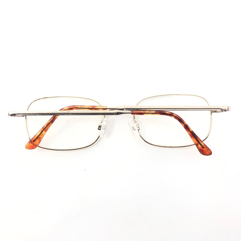 May include: Rimless spectacles with a gold-coloured metal frame and tortoiseshell patterned arms. The spectacles have a rectangular shape and clear lenses. The nose pads are clear. Designed for vision correction.