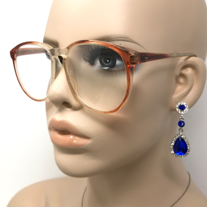May include: Clear-lens eyeglasses with a light brown frame are worn by a mannequin. Dangling earrings with a blue teardrop-shaped jewel and smaller blue and clear stones are also visible. The earrings are silver-toned.