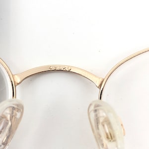 May include: Close-up of gold-colored eyeglasses with clear lenses. The bridge of the glasses has the number "49020" etched into the metal. The nose pads are clear plastic.