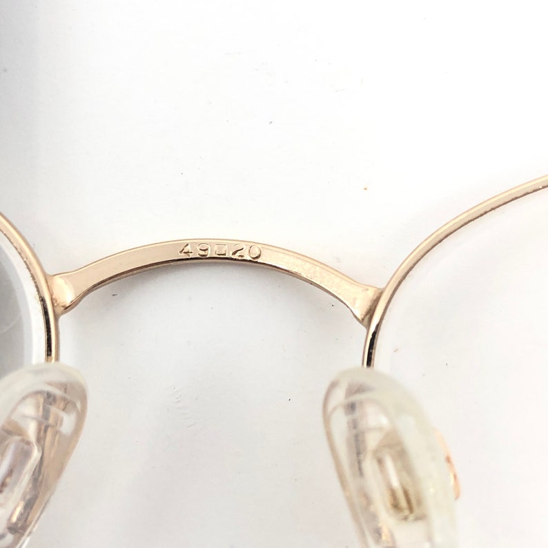 May include: Close-up of gold-colored eyeglasses with clear lenses. The bridge of the glasses has the number "49020" etched into the metal. The nose pads are clear plastic.